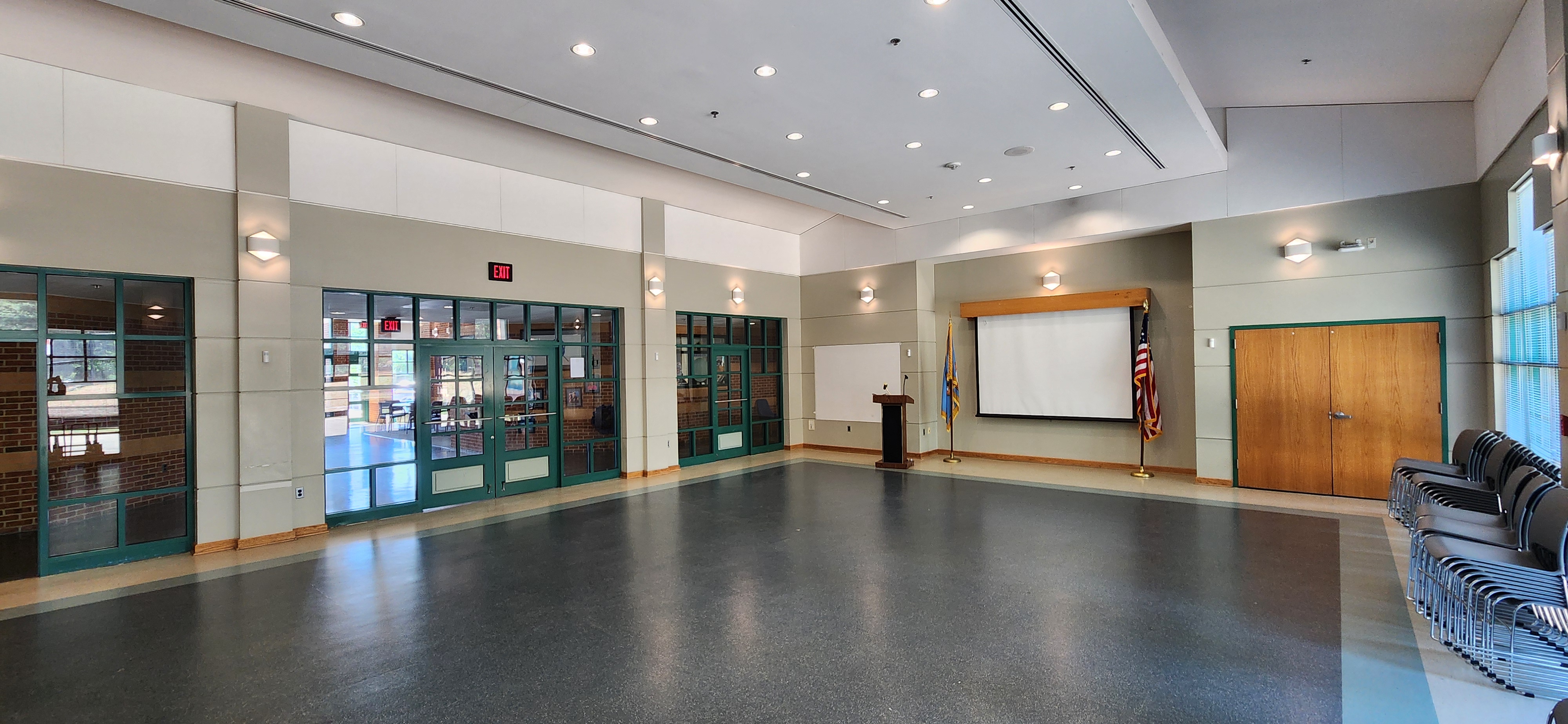 Spacious, bright room with a podium, projector screen, flags, green-framed glass doors, wood double doors, and stacked chairs by tall windows.