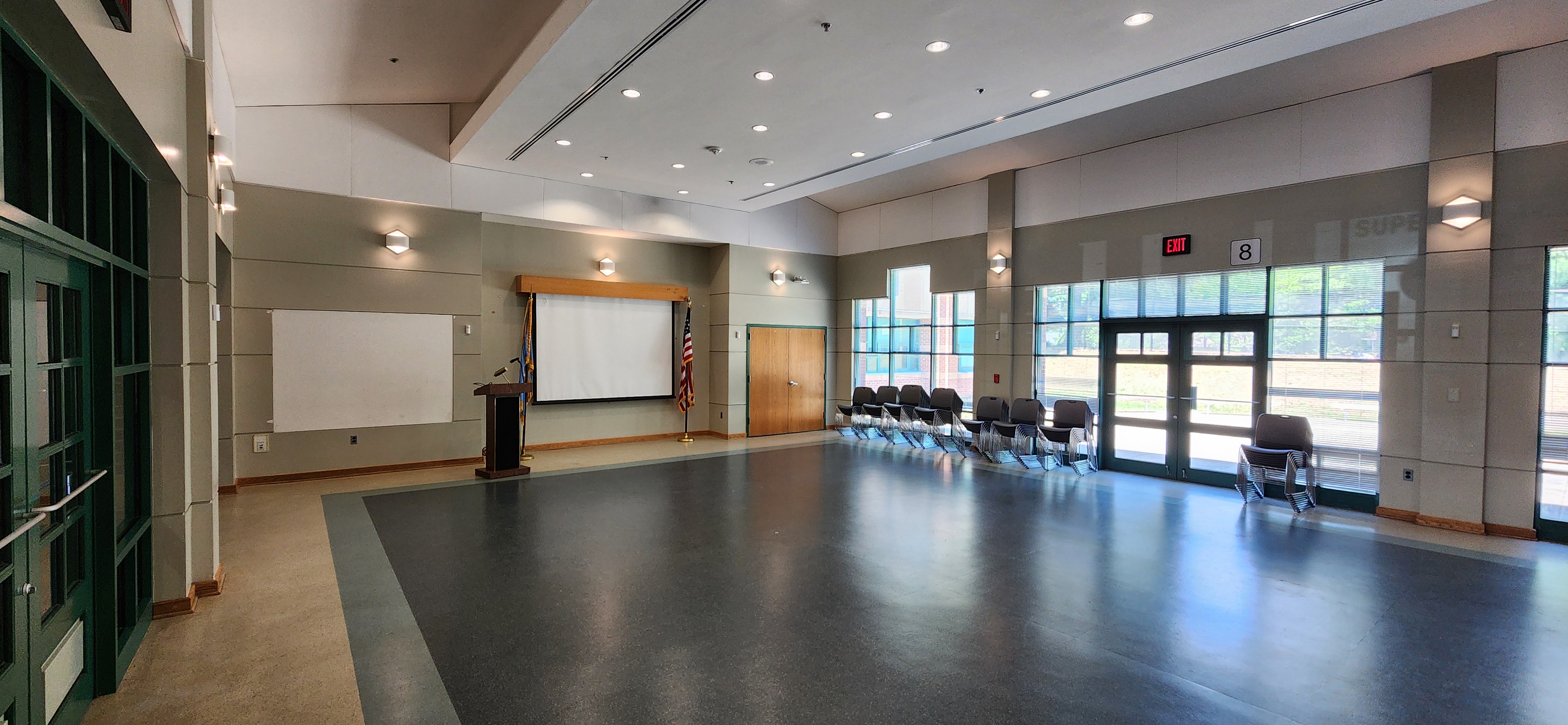Open room with a podium and screen at the front, stacked chairs along the right windows, and green-framed doors lining the left wall.