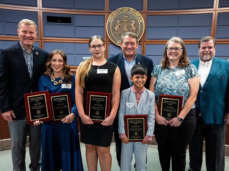 Adults in business attire stand next to youth holding plaques in an auditorium.