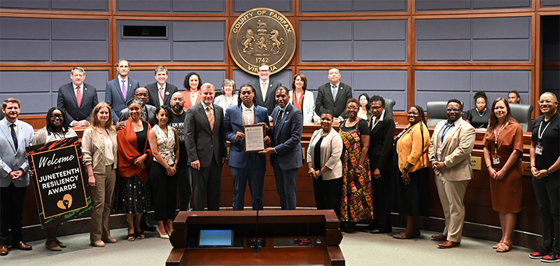 A group of individuals in business attire pose with a certificate in a government meeting room with the Seal of Fairfax County on the wall.
