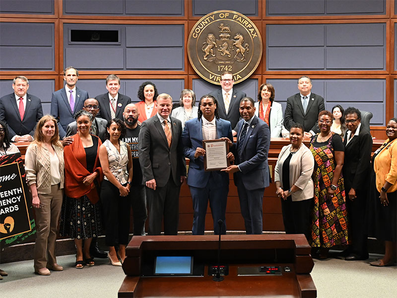 A group of individuals in business attire pose with a certificate in a government meeting room with the Seal of Fairfax County on the wall.