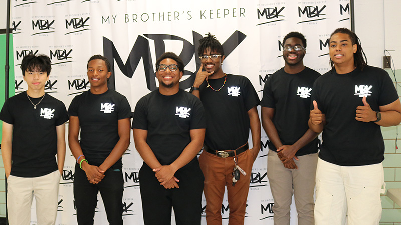 Young men in matching shirts in front of a step and repeat banner featuring the My Brother's Keeper logo.
