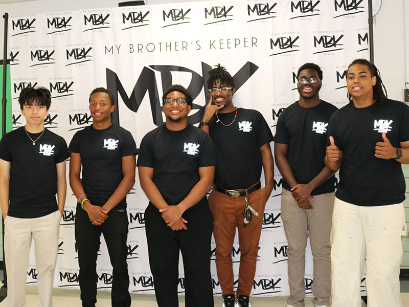 Young men in matching shirts in front of a step and repeat banner with the My Brother's Keeper logo.