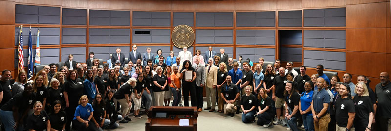 A group of individuals in business attire pose with a certificate in a government meeting room with the Seal of Fairfax County on the wall.