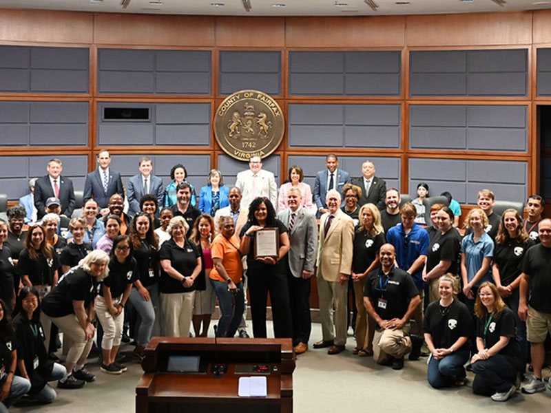 A group of individuals in business attire pose with a certificate in a government meeting room with the Seal of Fairfax County on the wall.