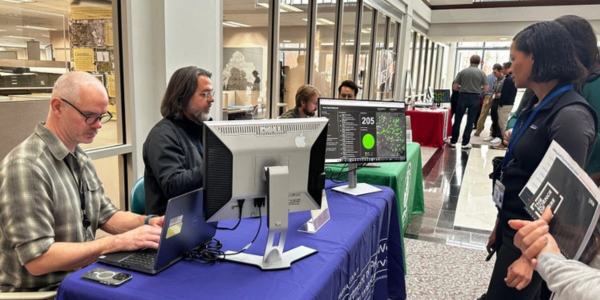 People gather at the 2024 GIS Day near a table with computer monitors and educational materials. Others engage nearby.