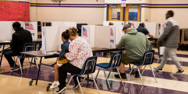 This image shows a polling station where individuals are seated at tables casting their votes. Each person is using a voting booth for privacy, with a white divider displaying an American flag and the word "VOTE."