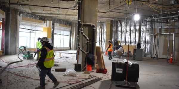 Interior of a large building under construction, with exposed concrete floors, open ceilings with wiring and pipes, and partially framed walls. Several workers wearing safety vests, hard hats, and protective gear are walking or operating equipment. Construction tools, cables, and materials are scattered across the floor, and bright work lights illuminate the space.