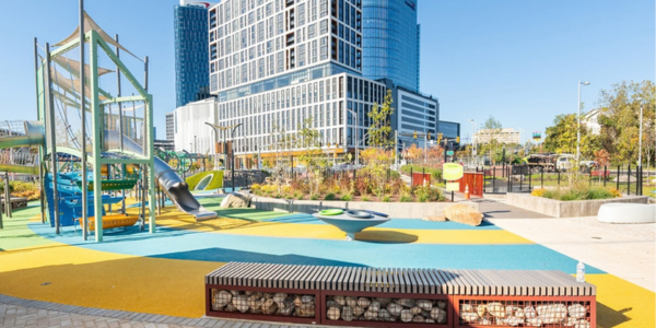 Colorful urban playground at Capital One East Park with modern play structures, landscaped rain gardens, and a high-rise office building in the background.