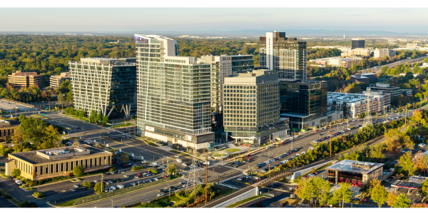 A wide aerial view of Reston, Virginia, showing a cluster of modern high‑rise office and residential buildings along a major multi‑lane roadway. The scene includes busy intersections, smaller commercial buildings, and tree‑filled areas extending into the distance under clear skies.