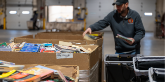 Worker in a warehouse sorting stacks of withdrawn library books in large cardboard bins