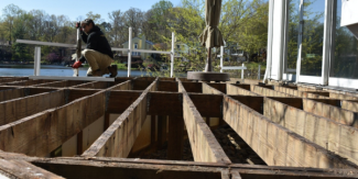Partially demolished wooden deck showing exposed joists, with a person kneeling on one of the beams and working. A lake, homes, and trees are visible in the background.