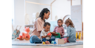An adult sits on a rug in a bright playroom with three young children, helping them explore toys and activity materials. The children are seated around the adult and are engaged with colorful objects, including blocks and a ball. The background shows shelves, large toys, and soft natural lighting.