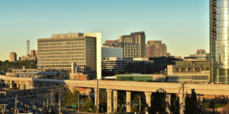 Skyline view of Tysons with several modern mid‑ and high‑rise office buildings, a Metro station, and an elevated rail line in the foreground, all under a clear sky.