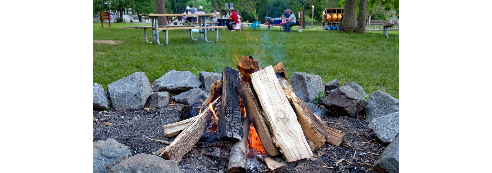 A campfire sits ready for lighting. Picnickers eat in the background.