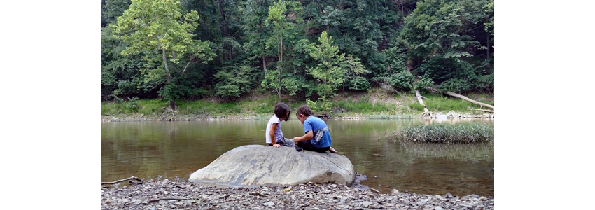Two children sit on a rock alongside a stream