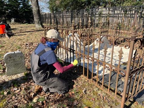 a CART staff members squats next to a rusty-looking fence around a some covered headstones, working to paint and stabilize the fence