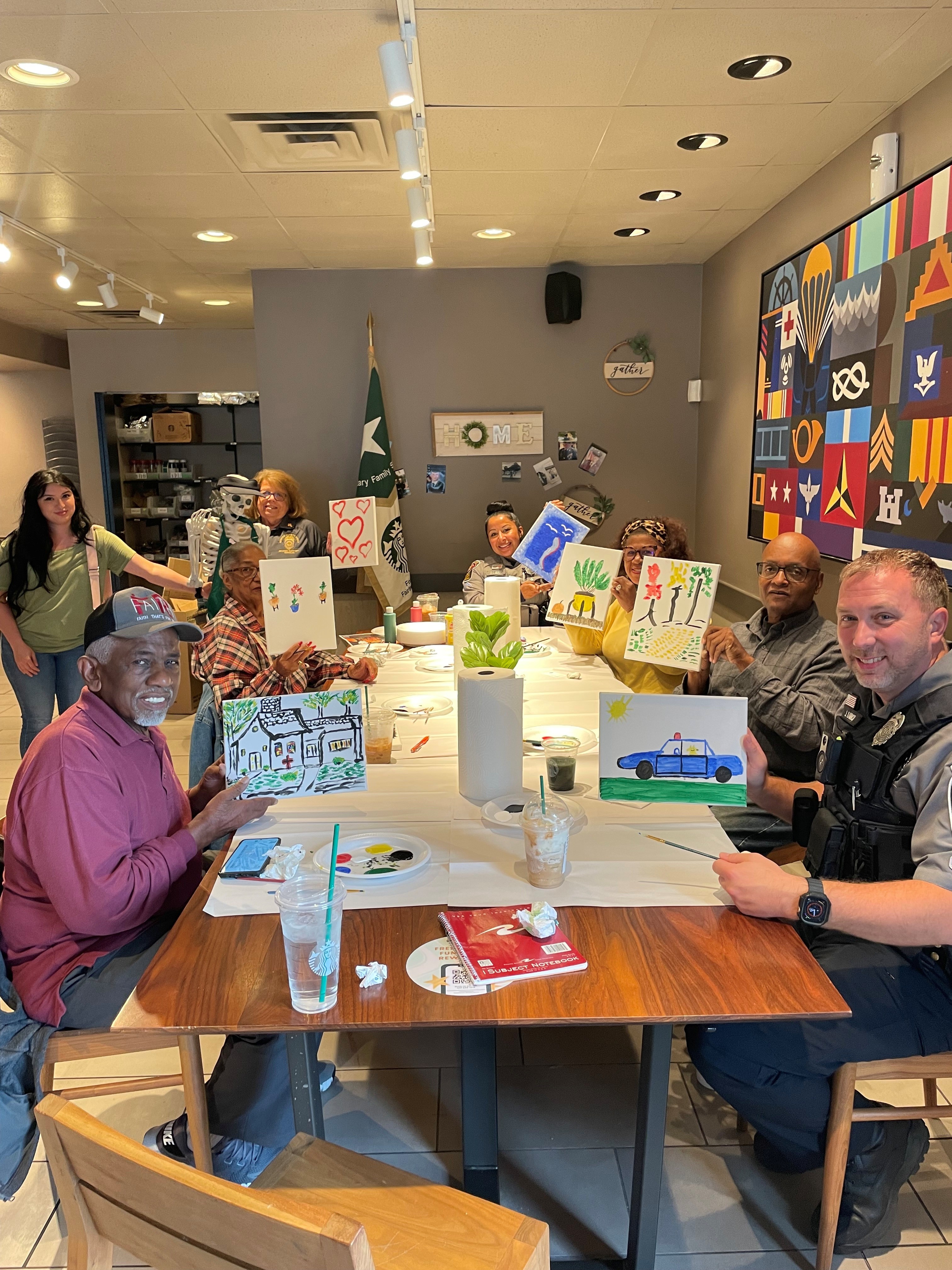Community members and a Fairfax County police officer sit around a table at a Faith & Blue paint event, holding up their finished paintings and smiling