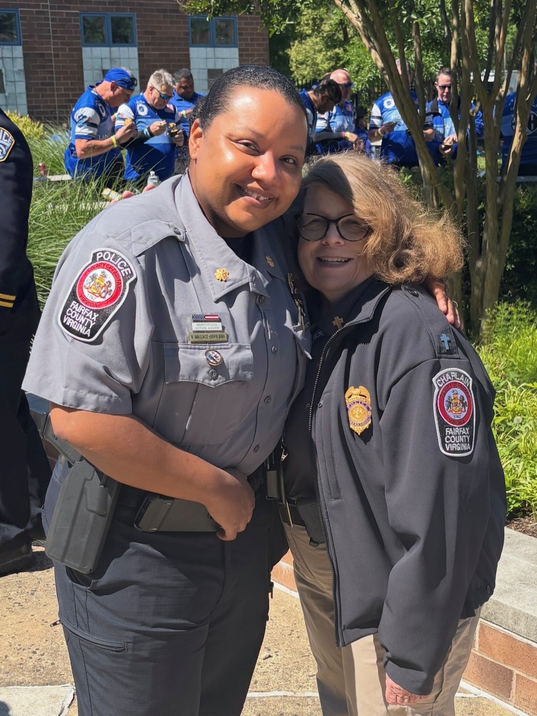 A Fairfax County police officer and a Fairfax County police chaplain embrace and smile at an outdoor Faith & Blue event