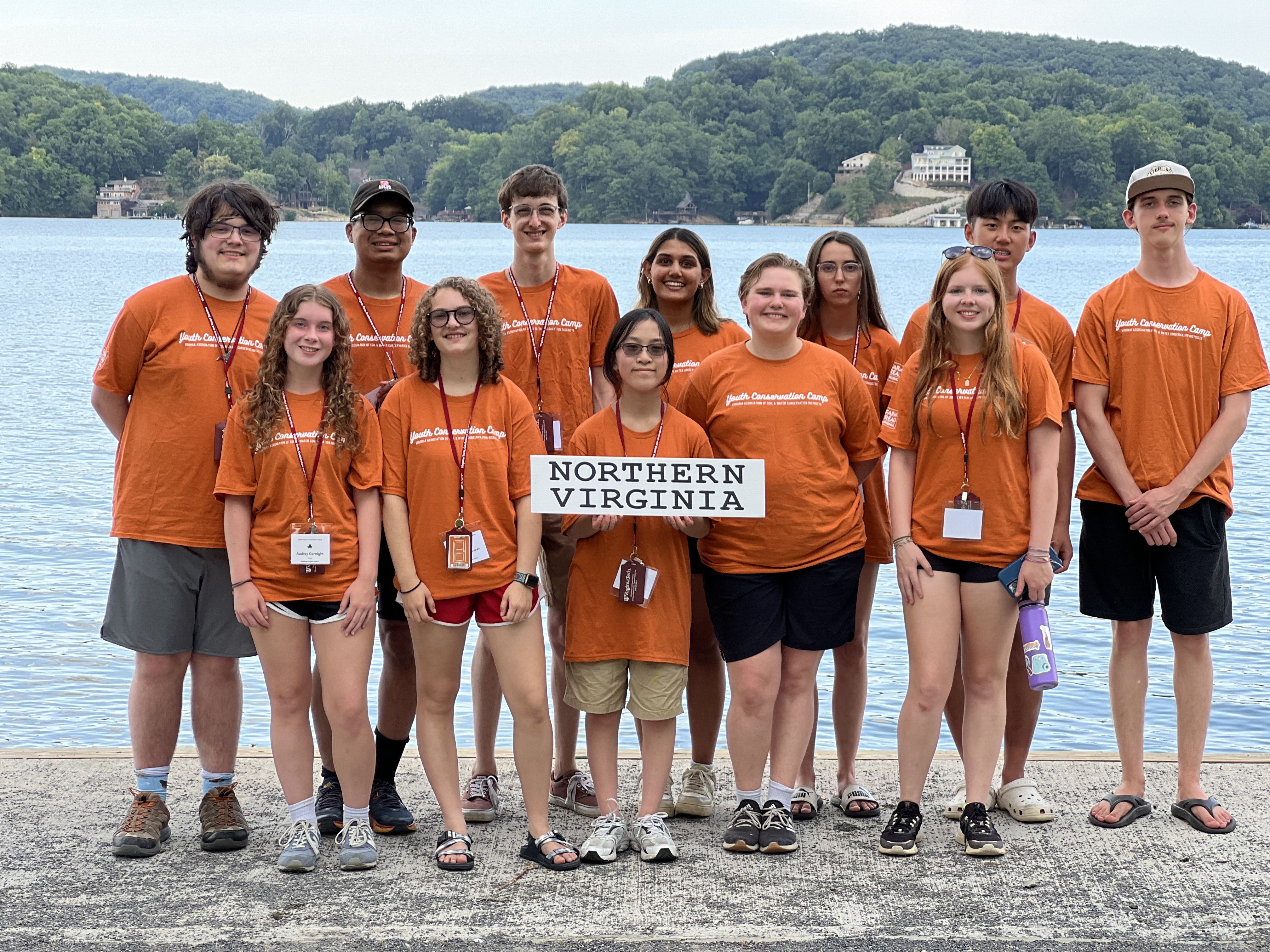 12 teenagers wearing orange shirts and lanyards stand in front of a lake with green hills behind it. The foremost camper holds a white sign that says "Northern Virginia" in black text. All of the students are smiling.